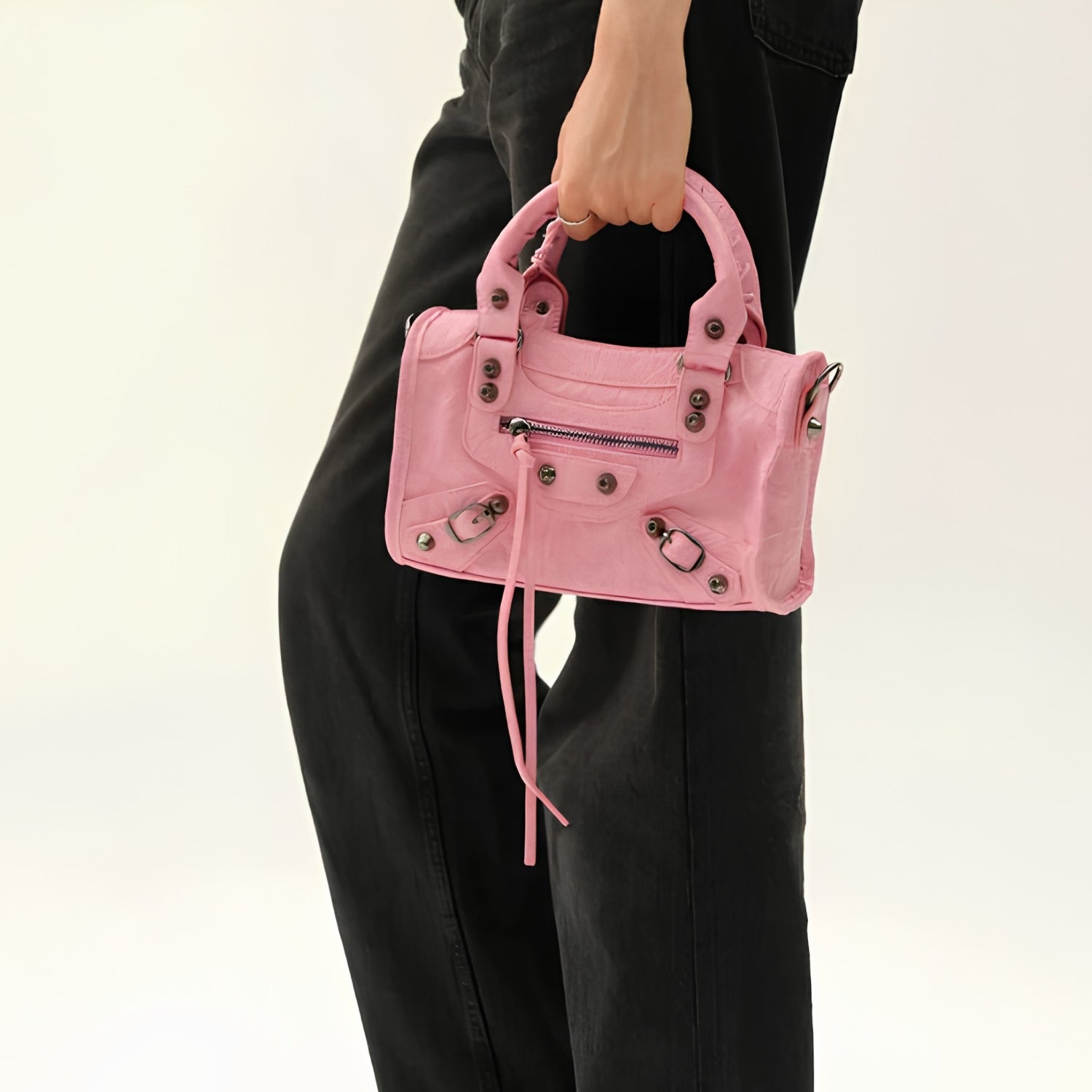 A person wearing black pants holds a small pink leather handbag with top handles, silver-tone hardware, and a front zip pocket against a plain light background.