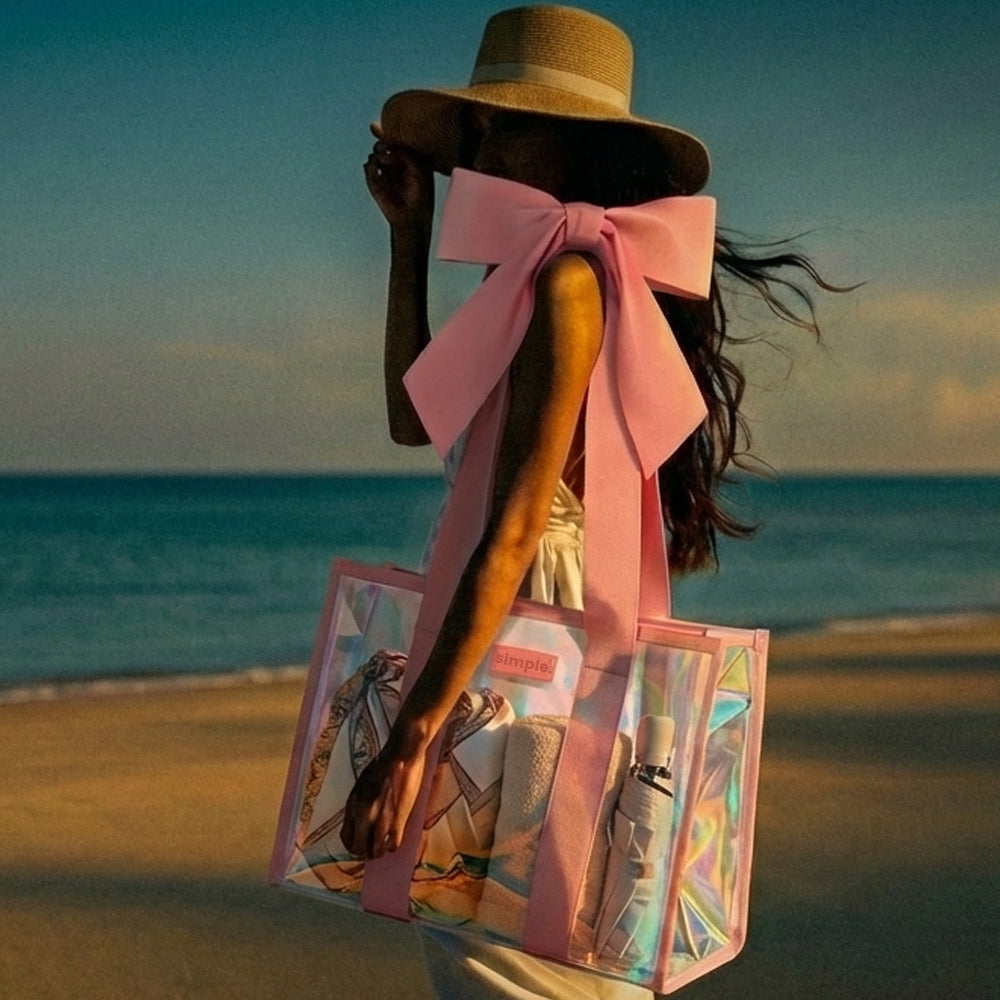 A woman on a beach carries a clear holographic tote bag with a large pink bow and pink trim. The scene is backlit by warm golden-hour light, with the ocean and sky in the background.