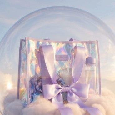 A pastel lavender and iridescent holographic gift bag with a large satin bow, containing a small bottle, set against a dreamy sky background with clouds.